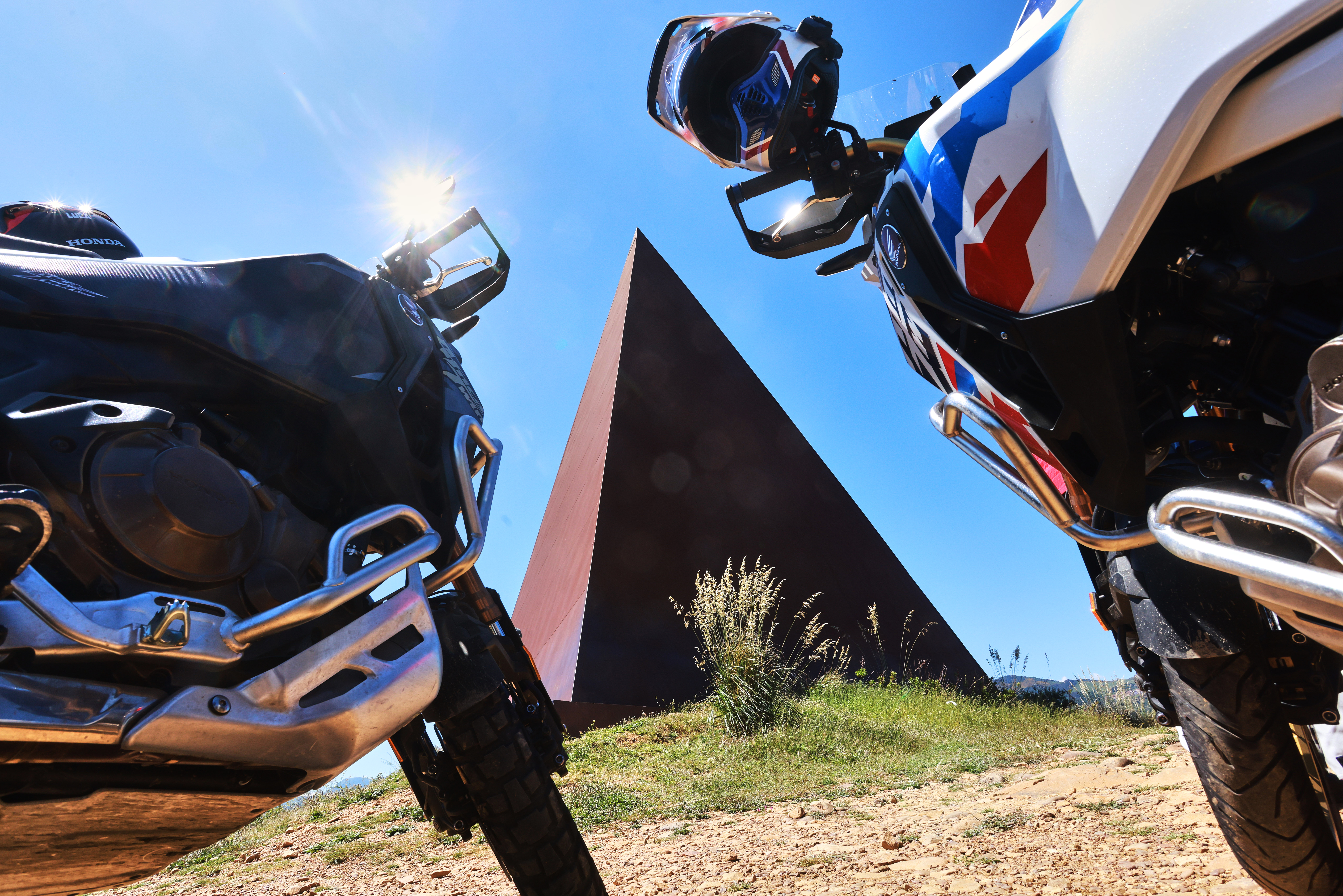 Due moto Honda Africa Twin posizionate davanti a una grande struttura a forma di piramide rossa sotto un cielo azzurro limpido.